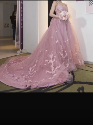 Rosa långklänning med spetsdetaljer - Magisk rosa långklänning med voluminös kjol i tyll och vackra blommiga spetsapplikationer. Klänningen har smala axelband och en elegant, romantisk look som passar perfekt till bal eller fest. Materialet är lätt och ger en drömmig känsla.