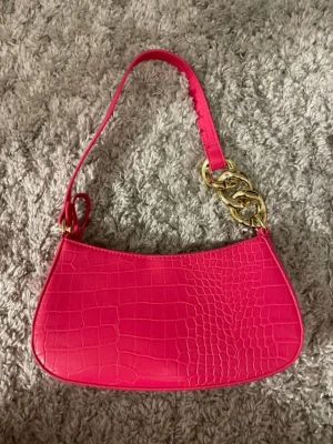 Rosa axelväska från Primark - Säljer en snygg rosa axelväska från Primark med krokodilpräglad yta och guldfärgade kedjedetaljer. Väskan har dragkedja upptill och en extra axelrem som kan fästas. Perfekt storlek för det viktigaste och riktigt trendig färg. Aldrig använd!