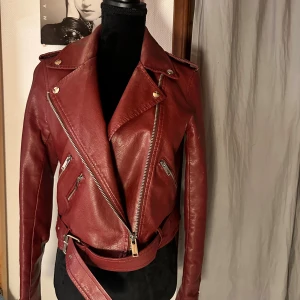 Vinröd skinnjacka från Fascinate - Säljer en snygg vinröd skinnjacka från Fascinate med coola silvriga dragkedjor, bred krage och bälte i midjan. Jackan har flera fickor med dragkedja och detaljerade axelpartier. Perfekt för dig som vill ha en edgy look.