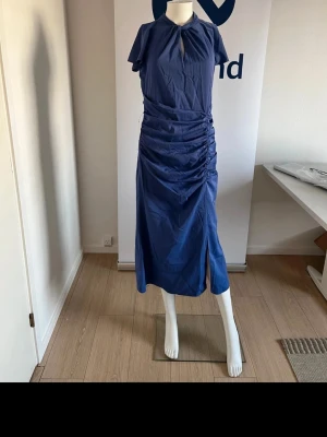 Lipsy London - Snygg blå långklänning från Lipsy med korta ärmar och draperad midja. Klänningen har en liten slits framtill och ett nyckelhålsformat öppet parti vid halsen. Materialet är mjukt och lätt, perfekt för dig som gillar stilrena detaljer och en modern siluett.