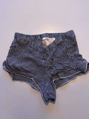 Randiga pyjamashorts från H&M - Säljer ett par korta, svartvita randiga shorts från H&M. De har elastisk midja och vita kanter längs benen. Perfekta för sommardagar eller som mjuka sovshorts. Materialet känns lätt och bekvämt.