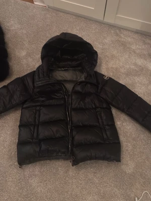 Svart pufferjacka från Colmar - Säljer en svart pufferjacka från Colmar med huva och dragkedja. Jackan har en quiltad look och Colmar-logga på ärmen. Perfekt för kalla dagar och riktigt snygg till streetwear-stilen.