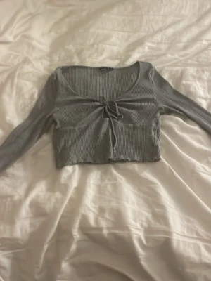 Grå ribbad croppad topp från SHEIN - Säljer en grå croppad topp från SHEIN i storlek M. Toppen har långa ärmar, ribbad struktur och en snygg knytning framtill vid bysten. Perfekt för att styla med höga jeans eller kjol. Materialet är mjukt och stretchigt syntet.den är använd några 2-3 gånger ingefär.