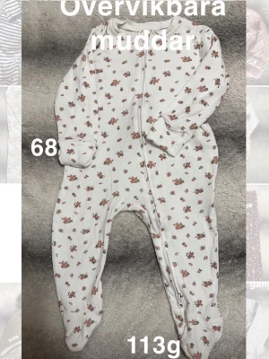 Blommig vit babypyjamas med dragkedja - Söt vit babypyjamas med små blommor i rosa. Pyjamas har dragkedja framtill och fötter samt övervikbara muddar på armarna. Perfekt för mysiga dagar hemma eller när du vill ha något gulligt och bekvämt till din lilla.🌸 Fint använt skick. Man kan vika över muddarna över händerna. Finns några mindre noppor. Storlek 68
