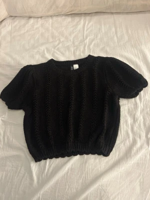 Svart stickad topp från H&M Divided - Svart stickad topp från H&M Divided i storlek S. Modellen har korta puffärmar och ett snyggt hålmönster i vertikala rader. Ribbad kant vid hals, ärmslut och nederkant. Perfekt att styla med jeans eller kjol för en chill look.