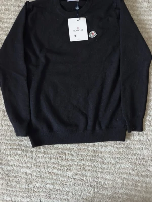 Svart Moncler stickad tröja - Svart stickad tröja från Moncler med rund hals och diskret Moncler-logga på bröstet. Tröjan har ribbade muddar vid ärmslut och nederkant samt långärmad passform. Perfekt för dig som gillar stilrena och klassiska plagg med exklusiv känsla.