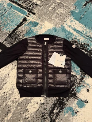 Moncler cardigan  - Säljer en fin stilig moncler cardigan i den klassiska glossiga svarta färgen som får jackan att sticka ut lite mer, perfekt för en enkel stilren look skriv om det är något du undrar 😊