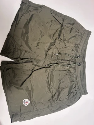 Olivgröna badbyxor från Moncler - Säljer ett par olivgröna badbyxor från Moncler med elastisk midja och snörning. Shortsen har meshfoder, en bakficka med dragkedja och Moncler-logga på benet. Perfekta för chill dagar eller när du vill ha en sportig look.