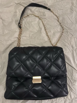 Svart quiltad axelväska med kedja - Säljer en svart quiltad axelväska med guldfärgad kedjerem och snyggt spänne framtill. Väskan är i mjukt skinnimitation och har ett stort huvudfack samt innerficka med dragkedja. Perfekt för dig som vill ha en stilren och trendig väska.