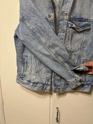 Ljusblå jeansjacka med knappar - Säljer en klassisk ljusblå jeansjacka från H&M med snygga metallknappar och två bröstfickor med lock. Jackan har en rak passform och långa ärmar. Perfekt att slänga på sig över en hoodie eller t-shirt för en chill look.