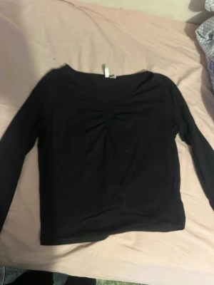Svart långärmad topp med rynk - En svart långärmad topp med snygg rynkdetalj framtill. Toppen har en enkel och stilren design med rundad halsringning och är gjord i mjukt material som känns skönt mot huden. Perfekt att matcha med jeans eller kjol.