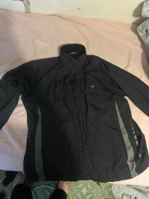 Svart vindjacka med reflexdetaljer - Säljer en svart vindjacka från Craft med grå reflexdetaljer längs sidorna. Jackan har hög krage, dragkedja framtill och diskret logga på bröstet. Perfekt för dig som vill ha en lätt och funktionell jacka till vardags eller träning.