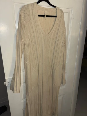 Beige ribbad lång stickad klänning kay/day - Säljer en beige lång stickad klänning från kay/day med v-ringning och långa ärmar. Klänningen har ribbat mönster, två stora fickor på sidorna och slits nertill. Perfekt för lager-på-lager och chill dagar.