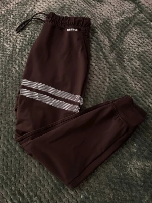 Svarta träningsbyxor från Stronger, storlek S - Svarta mjukisbyxor från Stronger med elastisk midja och snörning. Byxorna har två vita randiga detaljer på ena benet och är tillverkade i ett syntetmaterial som känns mjukt och följsamt. Passformen är normal och benen är smalare nedtill med muddar.