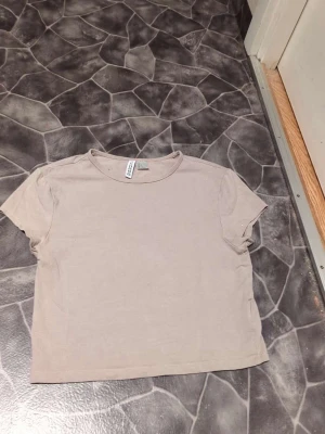Beige kortärmad t-shirt från H&M - En enkel och stilren beige t-shirt från H&M Basic. Modellen är kortärmad med rund hals och har en normal passform. Perfekt att matcha med jeans eller shorts för en chill look. Materialet är mjuk bomull som känns skönt mot huden.