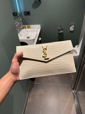 Beige kuvertväska från Saint Laurent - Säljer en beige kuvertväska från Saint Laurent i kalvskinn med krokopräglat mönster och guldigt YSL-emblem framtill. Väskan har magnetstängning och ett huvudfack. Helt oanvänd. Köpt för 670€ säljer endast för 4500kr. Priset kan diskuteras✅
