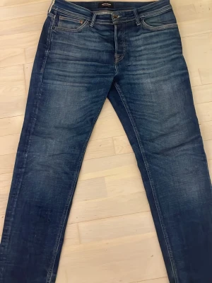 Jack & Jones Tapered Mike jeans blå - Snygga blå jeans från Jack & Jones, modell Tapered Mike. Jeansen har en klassisk femficksdesign, orangea sömmar och en cool tvättad look. Passformen är tapered vilket ger en modern siluett. Perfekta för dig som gillar stilrena och bekväma jeans.