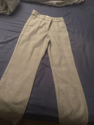 Beige rutiga kostymbyxor - Snygga beige kostymbyxor med diskret rutigt mönster och raka ben. Klassisk design med bälteshällor och knappstängning framtill. Perfekta för en stilren och trendig look.