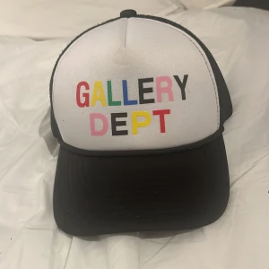 Truckerkeps från Gallery Dept. - Snygg truckerkeps från Gallery Dept. med vit front och svart mesh baktill. Framsidan har färgglad text i rött, gult, grönt, blått, svart och rosa. Justerbar snapback och broderade detaljer på insidan. Perfekt för en streetwear-look.