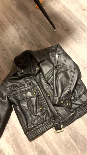 Svart skinnjacka med bälte - Cool svart skinnjacka med flera fickor, dragkedjor och snygga guldfärgade detaljer. Jackan har ett brett bälte i midjan och klassisk krage. Perfekt för dig som vill ha en edgy och tidlös look.