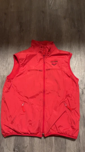 Röd västjacka från Mac One - Röd västjacka från Mac One med hög krage och hel dragkedja framtill. Jackan har två sidofickor med dragkedja och en bröstficka med dragkedja. Insidan är fodrad med mjuk fleece för extra komfort. Perfekt för lager-på-lager under kyliga dagar.