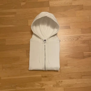 Vit zip-hoodie från Junkyard S - Vit hoodie från Junkyard i storlek S. Modellen har dragkedja hela vägen, två fickor fram, dragsko i huvan och ribbade muddar. Mjuk insida i fleeceliknande material. Enkel och clean look, perfekt till vintern! Tveka inte att ställa frågor!