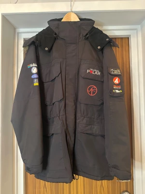 Svart parkajacka Fjällräven Polar, med i filmerna Van Veeteren - Parkajacka från Fjällräven Polar i svart med flera coola patches på ärmar och bröst. Jackan har varit med i filmserien Van Veeteren och har stor huva med fuskpäls, många fickor och tryck på ryggen. Perfekt för kalla dagar och har en riktigt schysst streetstil.