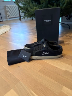 Saint Laurent sneakers - Letar du skor till jul? Perfekt! Säljer ett par svarta Saint Laurent sneakers med vit logga på sidan och svart snörning. Skorna har en beige platt sula och är tillverkade i mocka och skinn. Medföljer originalkartong och dustbag. Perfekt för dig som gillar stilrena och exklusiva sneakers. Skorna är endast använda ett par gånger och är även impregnerade!