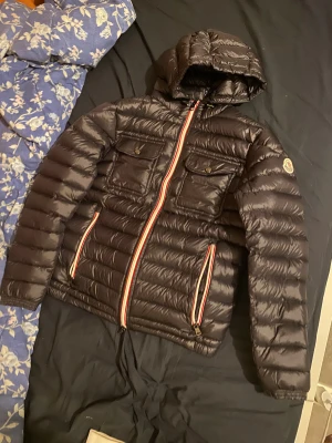 Svart Moncler dunjacka med huva - Svart dunjacka från Moncler med huva och coola röda, vita och blå detaljer vid dragkedjorna. Jackan har två bröstfickor med knappar och två sidofickor. Den är quiltad och har lång ärm, perfekt för kalla dagar. Snygg och klassisk look med Moncler-logga på ärmen.