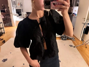 Svart blus med puffärm och knyt - Säljer en svart blus med puffiga korta ärmar och  knytningar framtill. Blusen har blommigt mönster i tyget och en lätt croppad passform. Knappt använd och super fin att ha på ny år❤️