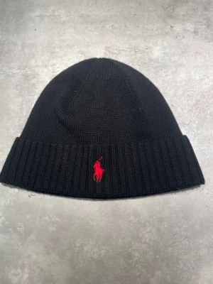 Svart mössa från Ralph Lauren - Svart mössa från Ralph Lauren med röd broderad logga framtill.  Klassisk design med uppvikt kant, perfekt för kalla dagar. 