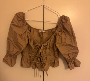 Beige blus med puffärm från NA-KD - Supersöt beige blus från NA-KD med puffiga trekvartsärmar och snygg snörning framtill. Croppad modell med smock i midjan och volangkant nedtill. Perfekt för dig som gillar romantisk och trendig stil.