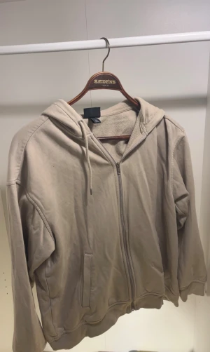 Beige hoodie med dragkedja från H&M - Snygg beige hoodie från H&M i loose fit med dragkedja framtill och två sidofickor. Mjuk fleece på insidan och klassisk huva med dragsko. Perfekt för chill dagar och enkel att matcha med jeans eller joggers.