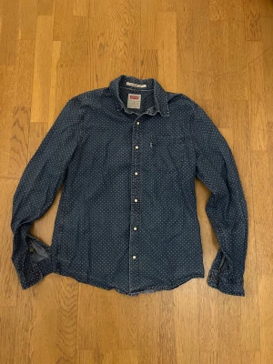 Blå prickig skjorta från Levi's - Snygg blå skjorta från Levi's med vita prickar och klassisk krage. Skjortan har lång ärm, bröstficka och knappar framtill. Modern fit som sitter skönt och passar till många olika stilar. Perfekt för dig som gillar coola detaljer och tidlös design.