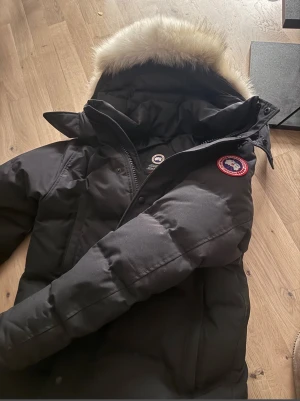 Canada goose Wyndham - Svart dunjacka från Canada Goose med röd logo. Jackan håller en riktigt varm under vintern och väldigt stiligt bland ungdomar. Kontakta gärna om ni har några frågor ☺️