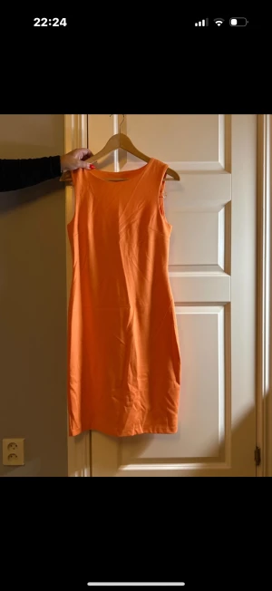 Orange ärmlös klänning med öppen rygg - Snygg orange klänning i enkel, rak modell med ärmlös design och cool öppen ryggdetalj. Perfekt för dig som gillar färg och vill sticka ut. Klänningen är knälång och har en stilren look utan mönster eller extra detaljer.