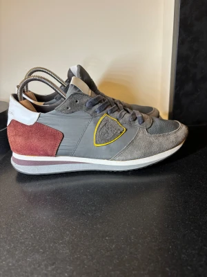 Philippe Model sneakers grå & vinröd - Snygga Philippe Model sneakers i grått med vinröda mockadetaljer på hälen och vita partier baktill. Skorna har mesh och mocka på ovandelen, snörning och en gul detalj runt loggan på sidan. Perfekta för dig som gillar stilrena men ändå unika sneakers.