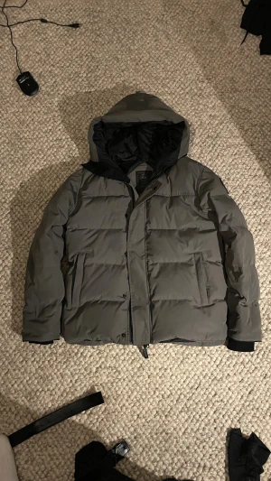 Grå pufferjacka från Canada Goose - Säljer en grå pufferjacka från Canada Goose i storlek M. Jackan har huva, svarta detaljer och ett snyggt Canada Goose-märke på ärmen. Fylld med dun och har dragkedja samt knappar framtill. Perfekt för kalla dagar och riktigt stilren look.
