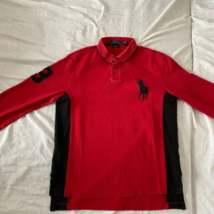 Röd pikétröja från Ralph Lauren - Säljer en snygg röd pikétröja från Ralph Lauren med svart broderad logga på bröstet och nummerdetalj på ärmen. Tröjan har en klassisk krage med knappar och långa ärmar. Perfekt för en stilren look!