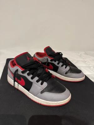 Nike Air Jordan 1 Low svart/röd/grå - Nike Air Jordan 1 Low sneakers med svart och grå ovandel i skinn, röda detaljer och röd yttersula. Klassisk rund tå, snörning och ikonisk Jumpman-logga på plösen. Snygg kontrast mellan färgerna och låg profil som passar streetwear-stilen.
