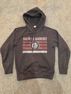 Mörkgrå Saint Laurent hoodie - Snygg mörkgrå hoodie från Saint Laurent med tryck i rött och vitt på bröstet. . Materialet är mjukt och bekvämt, perfekt för chill dagar. Trycket har en cool vintage-look med SL-emblem och stjärnor. Hoodien är givetvis äkta. Den är storlek M men sitter som S.