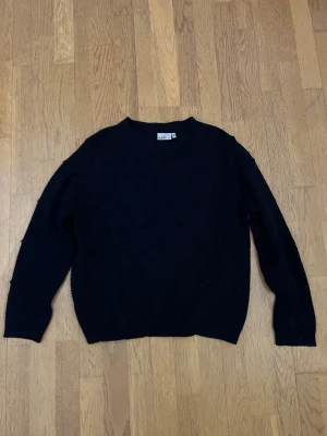 Svart stickad tröja från Isolde - Svart stickad tröja från Isolde i storlek M. Tröjan har rund halsringning och långärmade ärmar. Den är dekorerad med små diskreta bollar i stickningen som ger en cool textur. Perfekt för dig som gillar enkel men snygg stil.