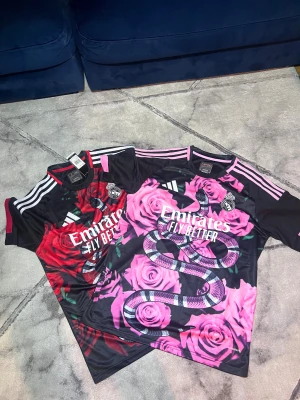 Real Madrid x Adidas blommig t-shirt - Säljer två Real Madrid t-shirts från Adidas med unikt blommigt mönster och ormtryck. Ena har rosa rosor och svarta detaljer, den andra röda rosor med svarta och gröna inslag. Båda har vita ränder på axlarna och tryckt klubbmärke på bröstet. Perfekt för dig som gillar statement-plagg.