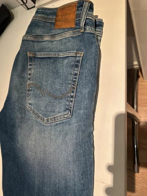 Blå jeans från Jack & Jones - Snygga blå jeans från Jack & Jones med klassisk femficksdesign och ljus tvätt. Jeansen har en skinnpatch med logga bak i midjan och kontrastsömmar. Perfekt passform och stilren look för dig som gillar casual denim.