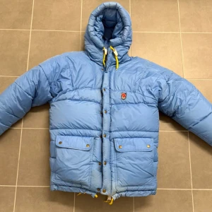Blå dunjacka från Fjällräven XS - Säljer en blå dunjacka från Fjällräven i storlek XS/S. Jackan har huva med gula snören, två stora fickor med knappar framtill och klassisk Fjällräven-logga på bröstet. Perfekt för kalla dagar med sin fluffiga och quiltade design. Sitter mer som S. 