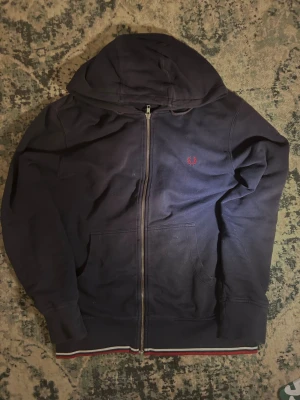 Mörkblå hoodie från Fred Perry M - Snygg mörkblå hoodie från Fred Perry med dragkedja och huva. Broderad röd logga på bröstet och ribbade muddar. Fickor framtill och cool randig detalj i rött, vitt och blått längst ner. Mjuk bomullskänsla, perfekt för chill dagar.