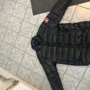 Svart dunjacka från Canada Goose - Säljer en svart dunjacka från Canada Goose med klassisk logga på ärmen. Jackan har quiltad design, dragkedja framtill och två sidofickor med dragkedja. Perfekt för kalla dagar och har en snygg, stilren look som funkar till det mesta. 