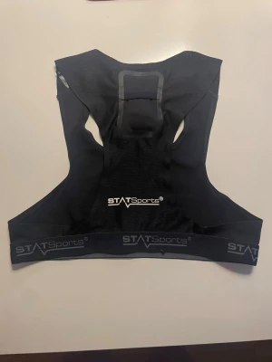 Svart träningsväst från STATSports - Svart träningsväst från STATSports i syntetmaterial, designad för att hålla en GPS-enhet på ryggen. Västen har elastiskt band med logotypen och meshpartier för ventilation. Perfekt för dig som vill tracka din prestation på fotbollsplanen.