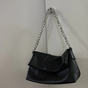 Svart axelväska med kedja - Snygg svart axelväska i skinnimitation med grov silverfärgad kedja som axelrem. Väskan har en mjuk, lite slouchy form 