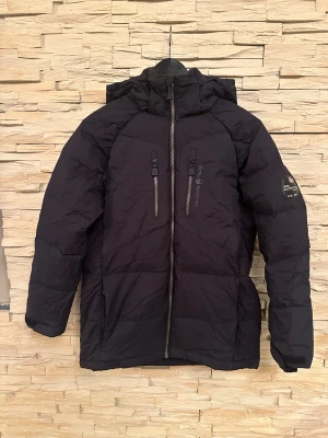 Svart dunjacka Sail Racing - Svart vinter jacka från Sail Racing med Sail Antarctica Expedition-märke på ärmen. Jackan har två bröstfickor med dragkedja, hög krage och huva. Jackan är perfekt för kalla vinterdagar. Snygg, sportig look med diskreta loggor och detaljer.